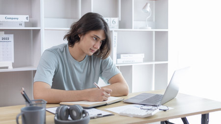 Asian man taking notes in notebook while studying online in laptop at home, Video chat, Online...