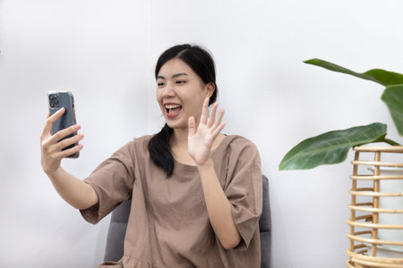 Young Asian woman happily using her mobile phone to vdo call  or talking with her friends, Entertaining or relaxing using mobile phone technology, Stay at home for long weekend, Happy lifestyle.の写真素材