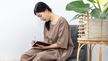 Asian woman sits in her living room writing a diary or notebook on the carpet, Weekends or holidays...