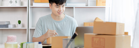 Young Asian man is writing down the customer's details and addresses on the notebook or box in order to prepare for shipping according to the information, Packing box, Sell online, freelance working.の写真素材
