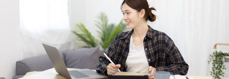 Asian woman taking notes in notebook while studying online in laptop or tablet  at home, Video chat, Online communication , Stay home, New normal, Distance learning
, Social distancing, Learn online.の写真素材