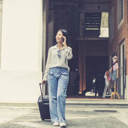 Asian woman holds a travel bag preparing to go to the accommodation and chat on the phone to a friend to meet up for a holiday party, Trolley bag, Long weekend travel, Tourism festival concept.の写真素材
