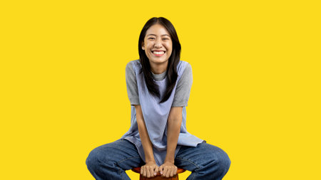 Asian biracial woman sitting in wooden chair and smiling happily on yellow background, Lovely lonely woman spreads a bright smile, Smile is cheerful and bright.の写真素材