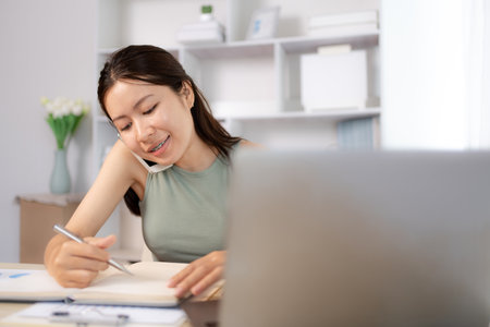 Young female college students talk on the phone and do homework at the same time, happy home life, Work from home.の写真素材