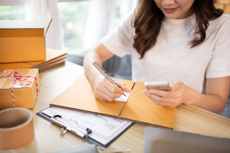 Woman Writing Customer Details for Shipping in Home Office, Packing Box and Writing Customer Information for Efficient Shipping, E-Commerce and Online Selling Concept.の写真素材