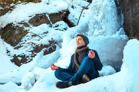 young man sitting in the snow snowdrift in the forest and looking at the winter riverの写真素材