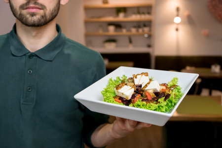 A waiter with a Greek salad on a white plate. Handsome young waiter at restaurantの写真素材