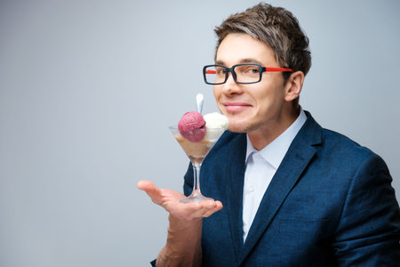 Handsome young man with glasses tastes ice cream balls in a transparent glass in the studioの写真素材