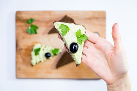 Top view of a woman holding a fresh avocado and making sandwiches on a table with other ingredients....