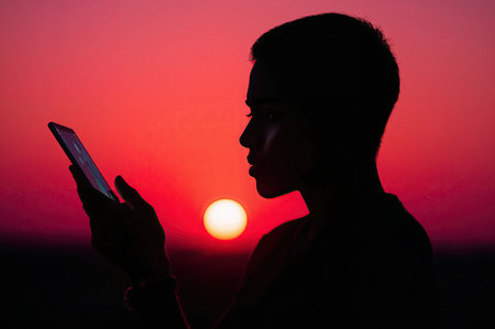 Young person silhouette reading tablet with sun on horizonの素材