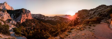 A sunset in the mountains. The sun at the bottom of the image shows us a landscape with a path and illuminated trees. In the background the mountains rise.の写真素材