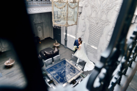 Wedding couple indoors is hugging each other. Beautiful model girl in white dress. Man in suit. Beauty bride with groom. Female and male portrait. Woman with lace veil. Cute lady and handsome guyの写真素材