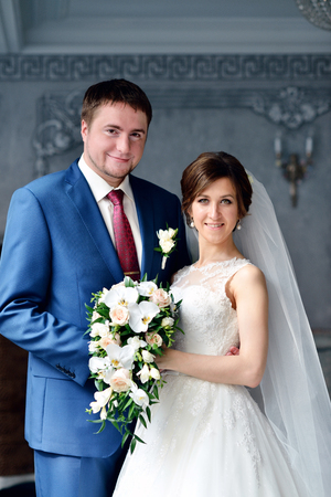 Wedding couple indoors is hugging each other. Beautiful model girl in white dress. Man in suit. Beauty bride with groom. Female and male portrait. Woman with lace veil. Cute lady and handsome guyの写真素材