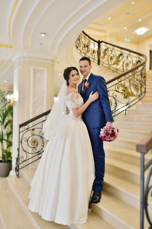 Wedding couple indoors is hugging each other. Beautiful model girl in white dress. Man in suit. Beauty bride with groom. Female and male portrait. Woman with lace veil. Cute lady and handsome guyの写真素材
