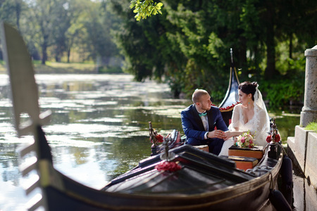 Wedding couple is hugging on a floating gondola. Beauty bride with groom. Beautiful model girl in white dress. Man in suit. Female and male portrait. Woman with lace veil. Cute lady and guy outdoorsの写真素材