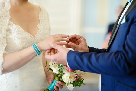 Beauty bride and handsome groom are wearing rings each other. Wedding couple on the marriage ceremony. Beautiful model girl in white dress. Man in suit. Female and male portrait. Cute lady and guyの写真素材
