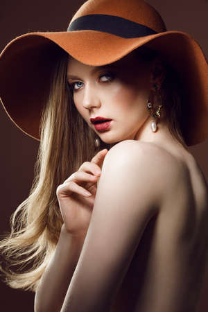 Fashionable female portrait of cute lady indoors with hat. Close up beautiful sexy model girl in elegant pose isolated on brown background. Closeup beauty blonde woman with hairstyle and makeupの写真素材