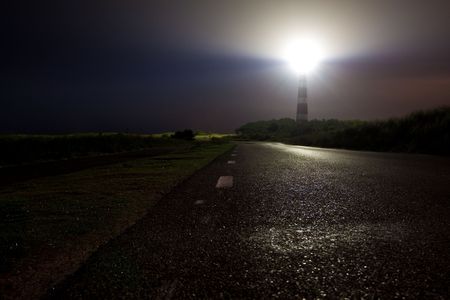 Lighthouse shining his light in the night seen from the roadの写真素材