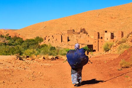 Moroccan woman carying a big bag with herbs with kasbah in the background against the hillsの写真素材