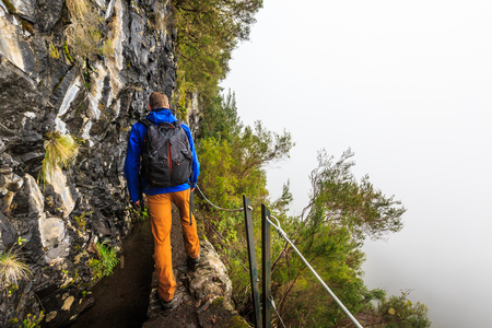 Man walking a levada trail with Steep abyss next to the trail in the mountains of Madeira, Portugal on a rainy misty dayの写真素材