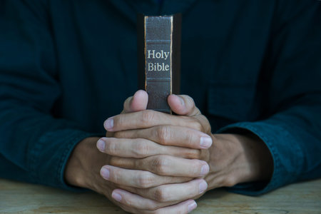 Christian man holding bible. Hands folded in prayer on a Holy Bible on wooden table.
