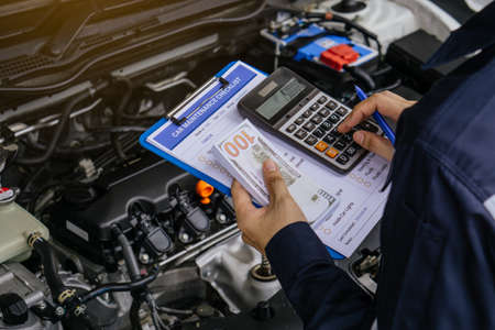 Hands of machinic man holding calculator and money, dollar banknotes to charge service and writing to the clipboard the checklist for repair machine, car service and maintenance.Services car engine machine concept.の写真素材