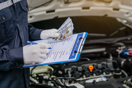 Hands of machinic man holding money, dollar banknotes and writing to the clipboard the checklist for repair machine, car service and maintenance.Services car engine machine concept.の写真素材