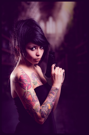Beautiful tattooed girl in black dress with attitude pointing guns up selective focusの写真素材