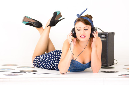 Beautiful young girl with headphones in retro clothes and hairstyle representing pinup style lying on the floor with vynil records isolated on whiteの写真素材