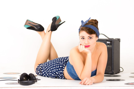 Beautiful young girl with hand under her chin in retro clothes and hairstyle representing pinup style lying on the floor with vynil records isolated on whiteの写真素材