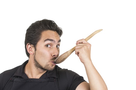 handsome young male chef wearing black apron tasting from wooden spoon isolated on whiteの写真素材
