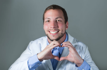 Closeup portrait of handsome young smiling doctor making a heart with hands over gray background