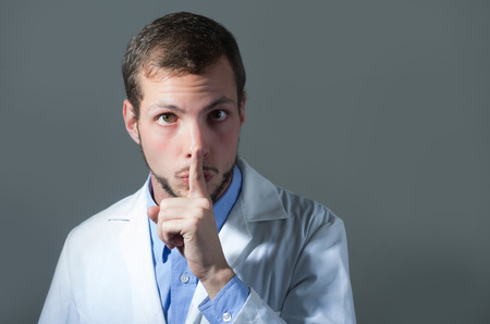 Closeup portrait of handsome young doctor gesturing shhh over gray backgroundの写真素材