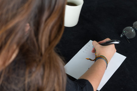 closeup of right hand writing on paper next to coffee mug and glasses with brown hair visible in...