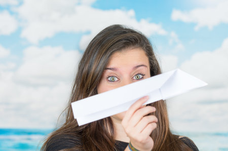 brunette holding a paperplane covering her nose in front of a cloud themed background ready to throw...