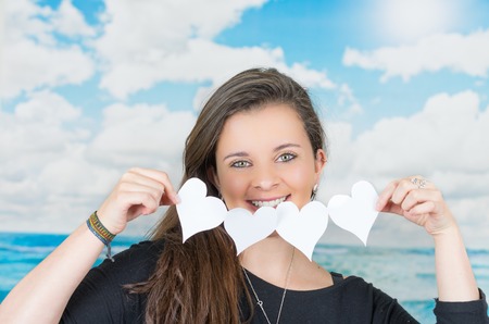 brunette holding an origami heartshaped paper figure in front of oceanic cloud background and smiling to cameraの写真素材