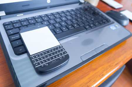 Open black laptop with black mobile phone placed on top of keyboard and wooden table background