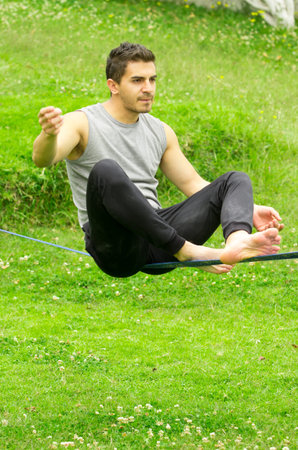 Man sitting on tightrope or slackline concentrating to keep balance with grassy background.