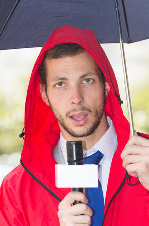 Successful handsome male journalist in red rain jacket working in rainy weather outdoors in park environment holding microphone and umbrella in live broadcasting.の写真素材