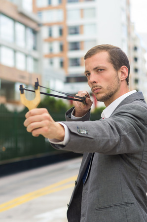 Handsome buisness man concentrated aiming a slingshot at urban city background.の写真素材
