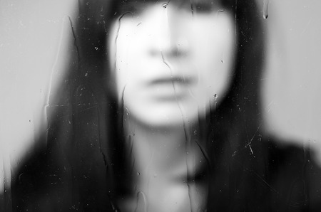 Black and white closeup diffused portrait of brunette girl behind glass concept of melancholy, sadness, lonelinessの写真素材