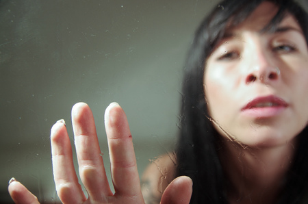 Beautiful sad young girl behind wet glass reaching out with her handの写真素材