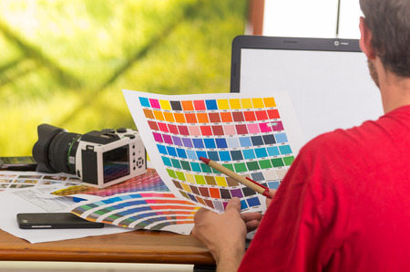 Man working on laptop while holding up pantone palette, colormap from behind angle. Concept graphic designer, renovation, color samples, interior design, printing industryの写真素材