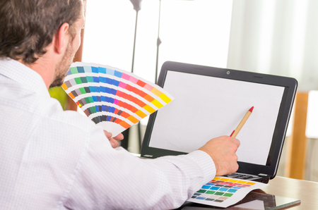 Man working on laptop while holding up pantone palette, colormap from behind angle.  Concept graphic designer, renovation, color samples, interior design, printing industryの写真素材