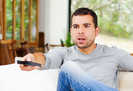 Hispanic male wearing light blue sweater plus denim jeans sitting in white sofa holding potato chip...