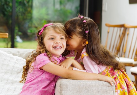Brunette children sisters sitting happily on white livingroom sofa posing for camera with typical pink girl clothes.の写真素材