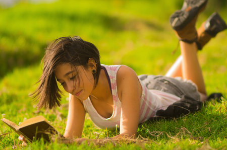 Pretty teenage hispanic girl wearing white top and shorts lying on grass relaxed reading from book.の写真素材
