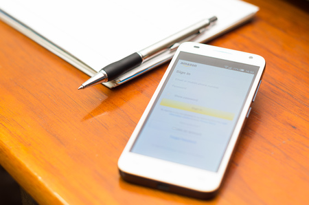 QUITO, ECUADOR - AUGUST 3, 2015: White smartphone lying on wooden desk with Amazon website screen next to a pen and notepad, business communication concept.のeditorial素材