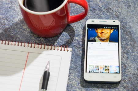QUITO, ECUADOR - AUGUST 3, 2015: White smartphone lying on table with Facebook website screen open next to a pen, notepad and coffee mug, business communication concept.のeditorial素材