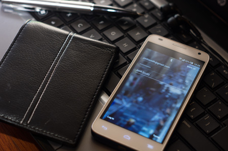 QUITO, ECUADOR - AUGUST 3, 2015: White smartphone closeup lying next to silver pen and wallet on laptop keyboard with unknown website login screen visible.のeditorial素材